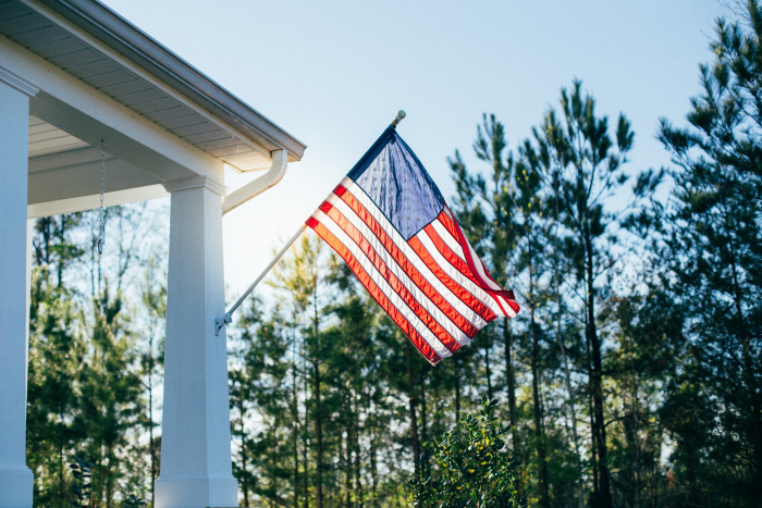 American flag waving outside porch in front of pine trees, backlit by the sun